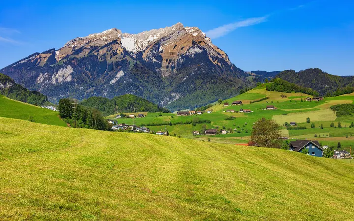 Plavo nebo i oblaci nad alpskim planinama s poljima i zgradama u podnožju.