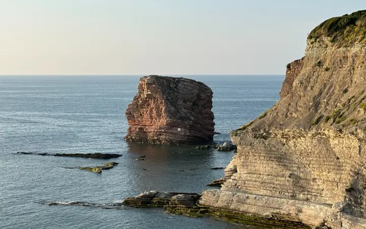 Plaža i litice uz obalu s plavim morem i svijetlim oblačnim nebom u prirodnom okruženju.