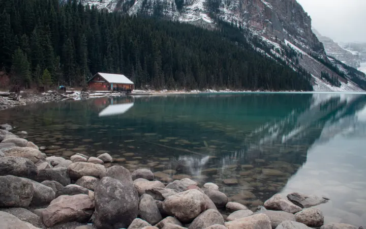 Mirno jezero okruženo oblacima i plavim nebom uz drvenu kućicu u prirodi.