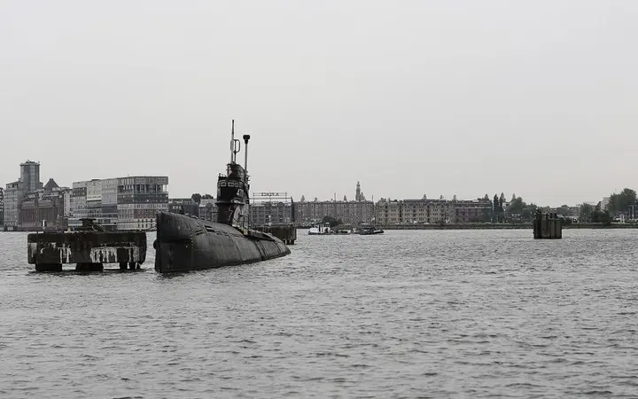 Podmornica smještena među brodovima i zgradama u gradskoj luci Amsterdama.