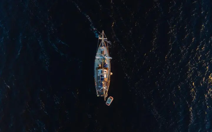 Pogled iz zraka na jedrilicu koja plovi tamnim oceanom, s prostranim plavim morem i otvorenim nebom.