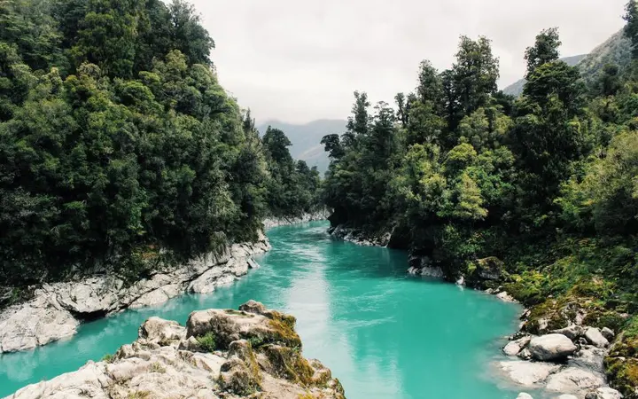 Zelena dolina okružena visokim planinama s rijekom u prvom planu i oblačnim nebom.