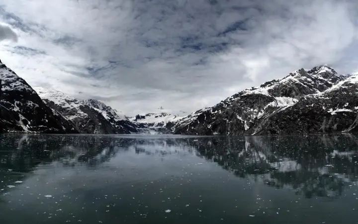 Mirno jezero okruženo planinama i oblacima s refleksijama u vodi u plavim i zelenim nijansama.